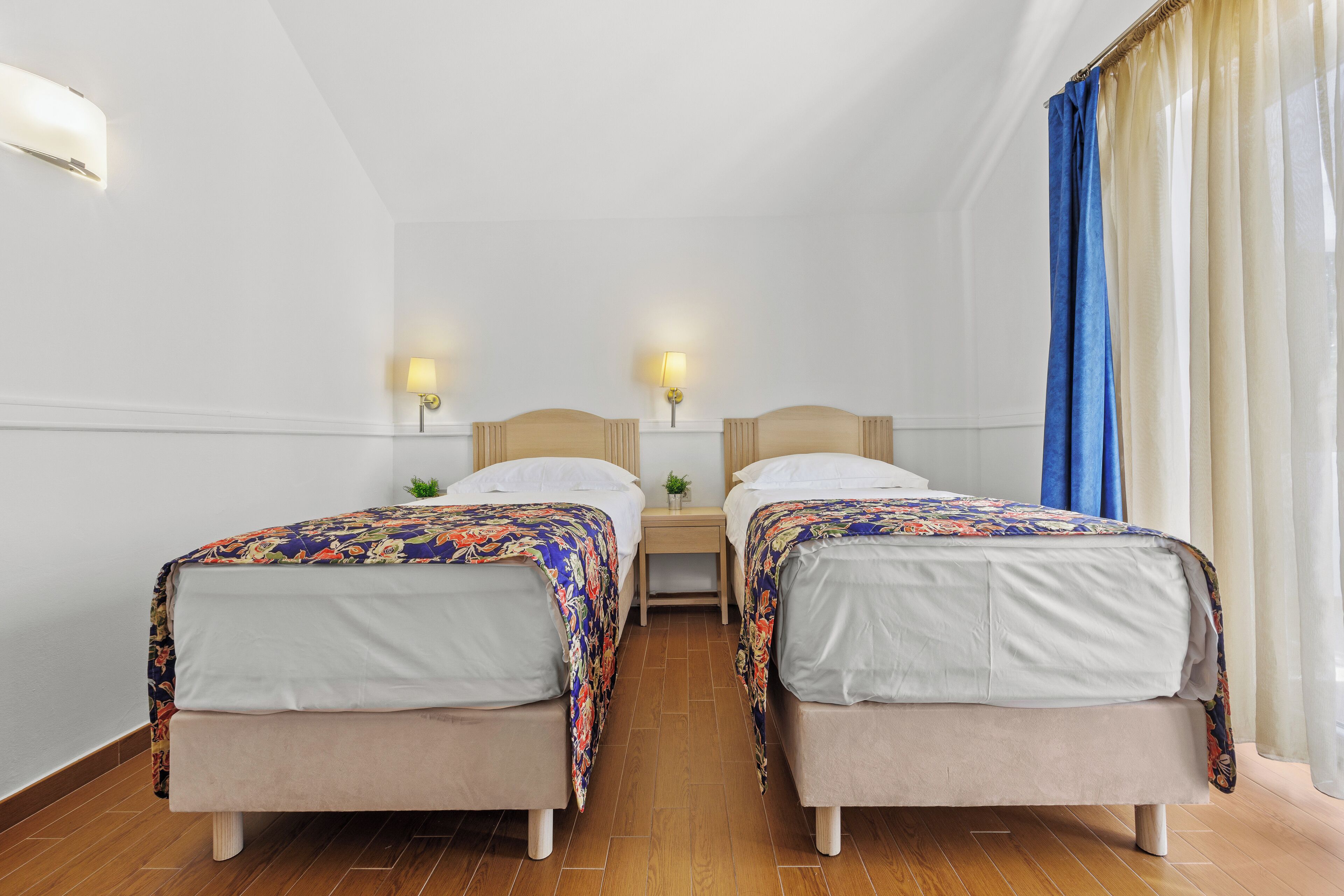 A hotel room with two single beds, a small bedside table with a lamp, and a window with curtains.