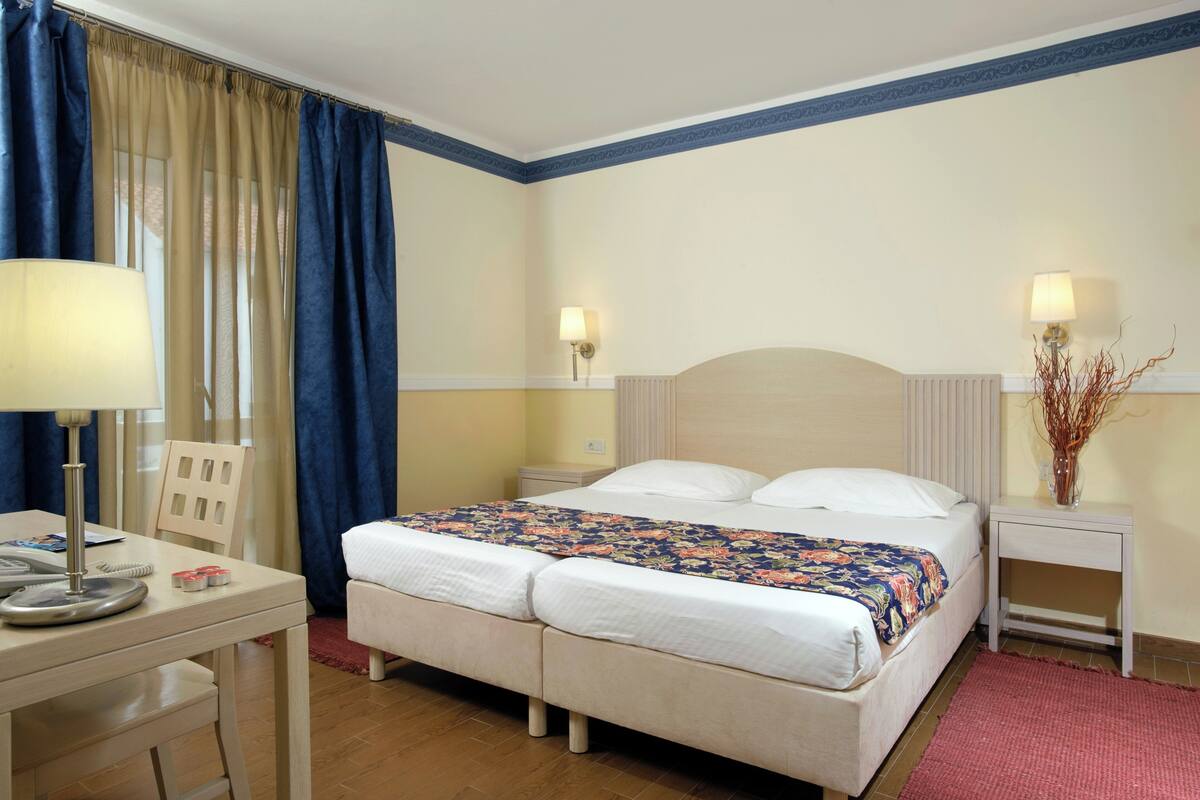 Habitación de hotel con cama doble, dos mesitas de noche, escritorio con lámpara, una silla y ventana con cortinas.