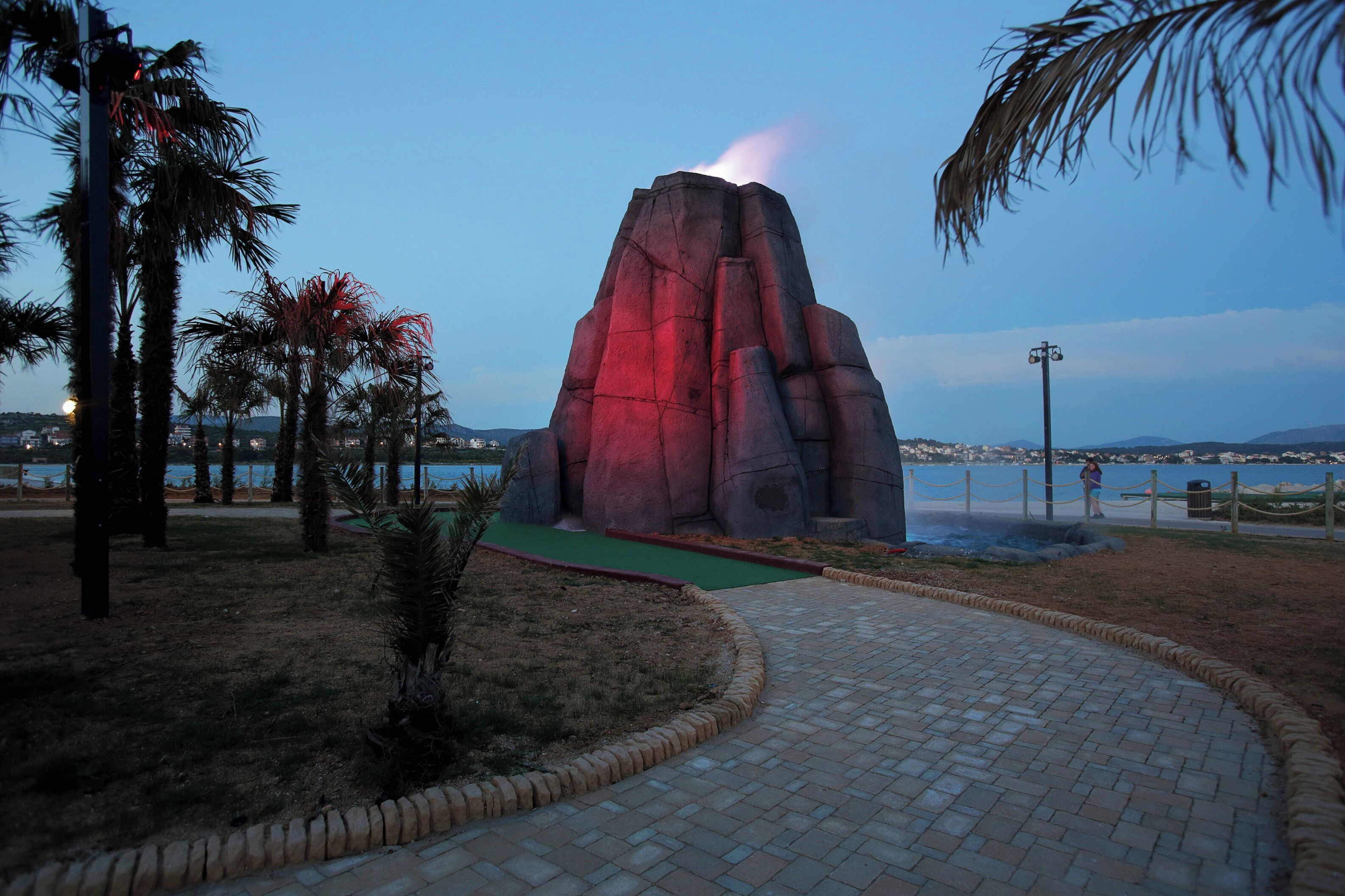 A landscaped area with a rock formation, palm trees, and a paved walkway.