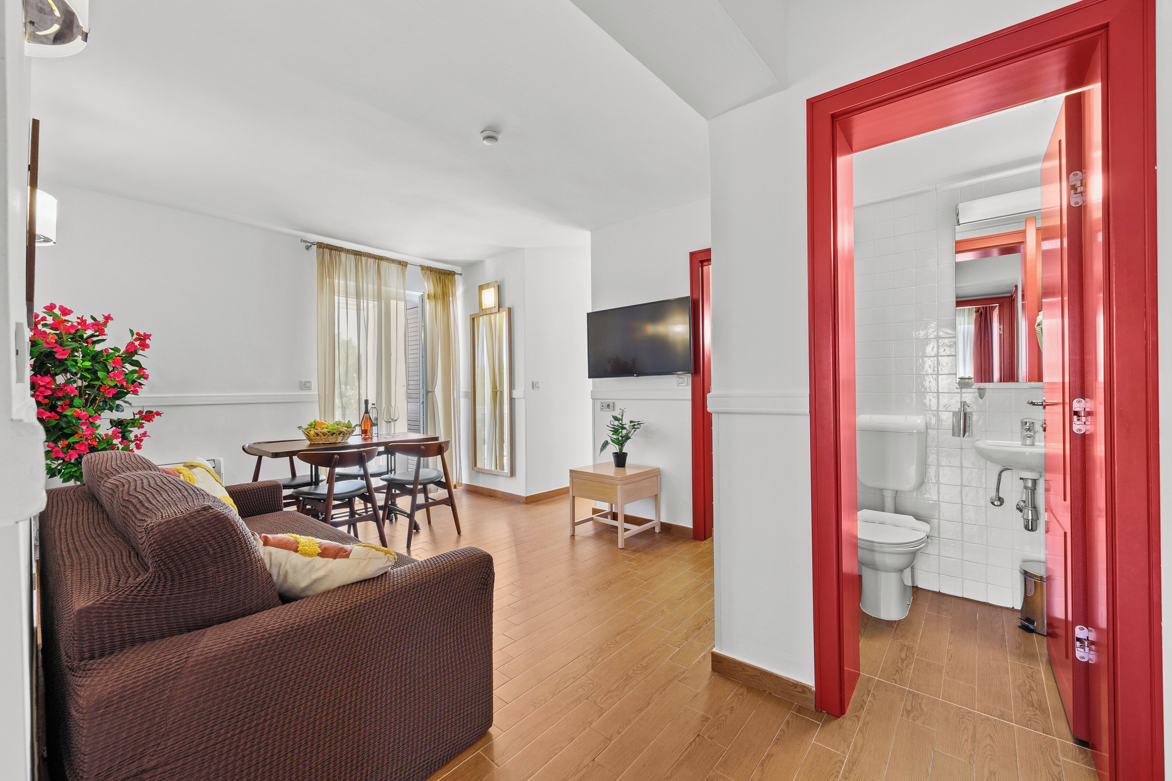A modern living room with a sofa, dining table, and a small side table. A TV is mounted on the wall, and a red door leads to a bathroom.