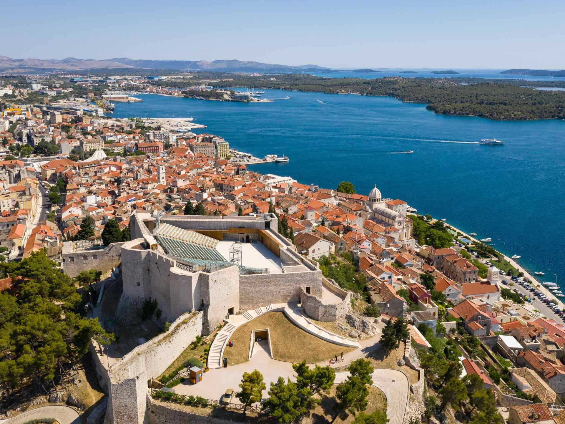 A coastal city with a prominent fortress, a marina, and a densely built urban area with red-roofed buildings.