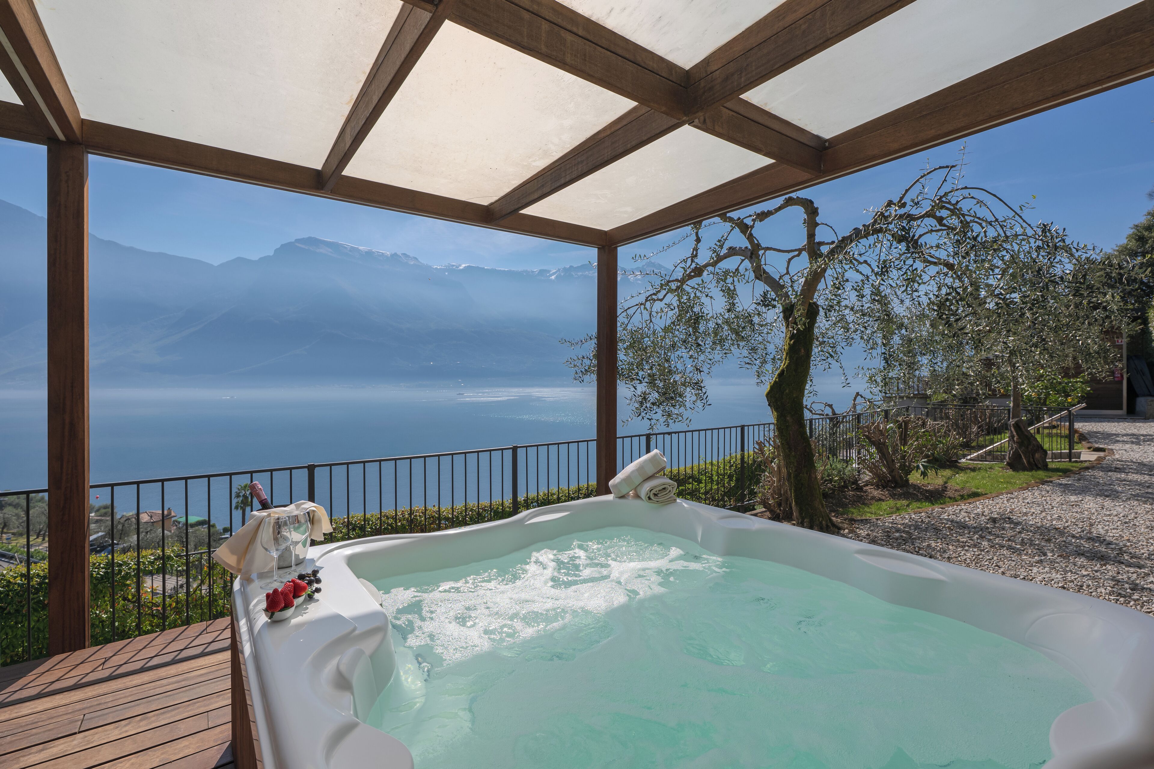 Un jacuzzi con vistas a un lago y montañas, rodeado por una terraza de madera y una barandilla metálica.