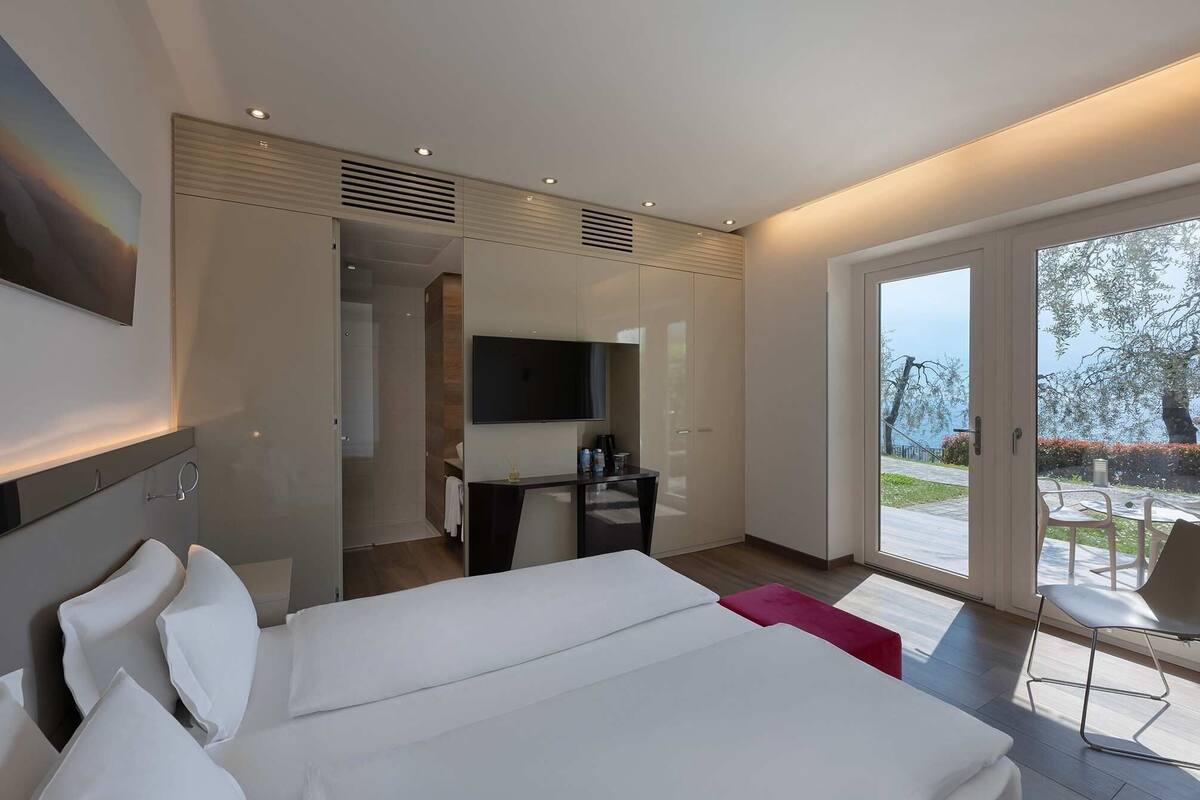 Ein modernes Hotelzimmer mit einem großen Bett, einem Fernseher, einem Schreibtisch und einem Balkon mit Sitzgelegenheit.