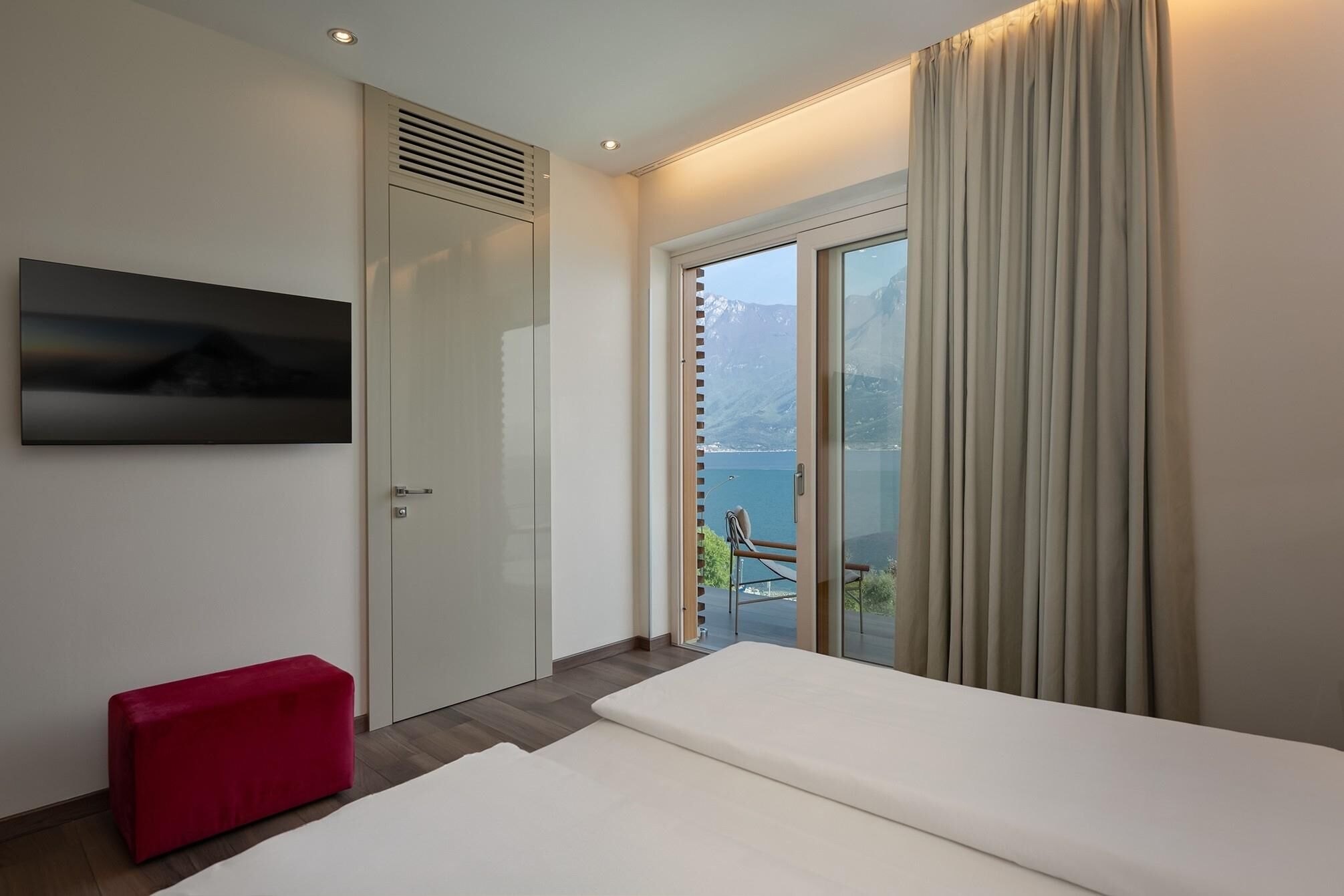 Habitación de hotel moderna con una cama, un banco rojo, un televisor y un balcón con vistas al mar y a las montañas.