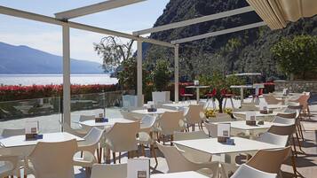 Una terraza con mesas y sillas, con vista a un cuerpo de agua y montañas.