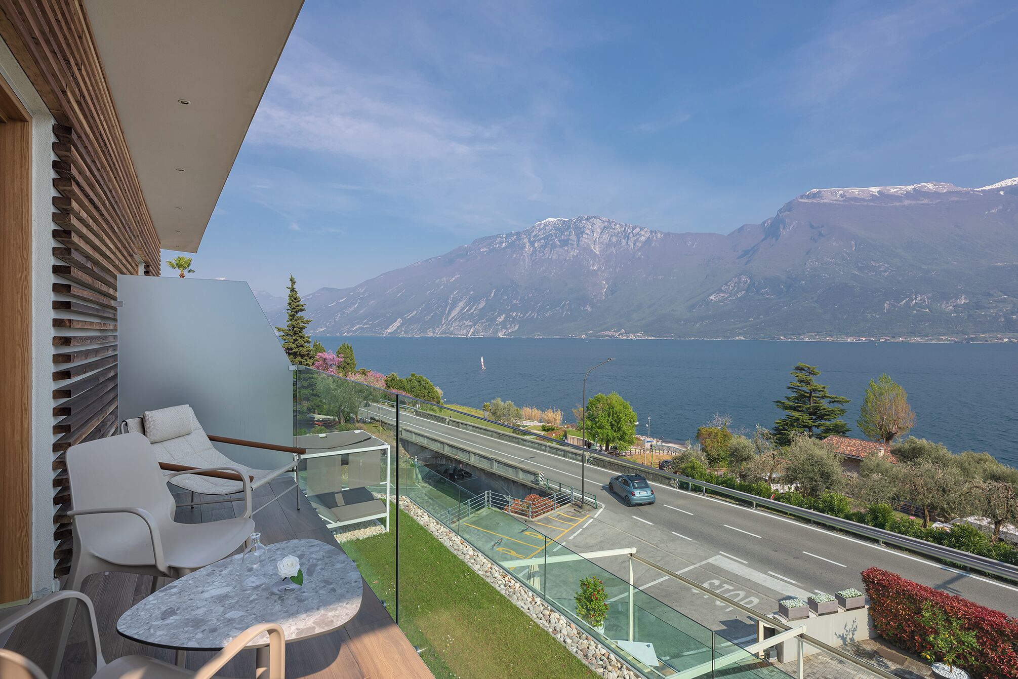A balcony with a view of a lake and mountains, a glass railing, and outdoor seating.