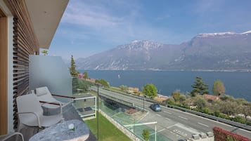 A balcony with a view of a lake and mountains, a glass railing, and outdoor seating.