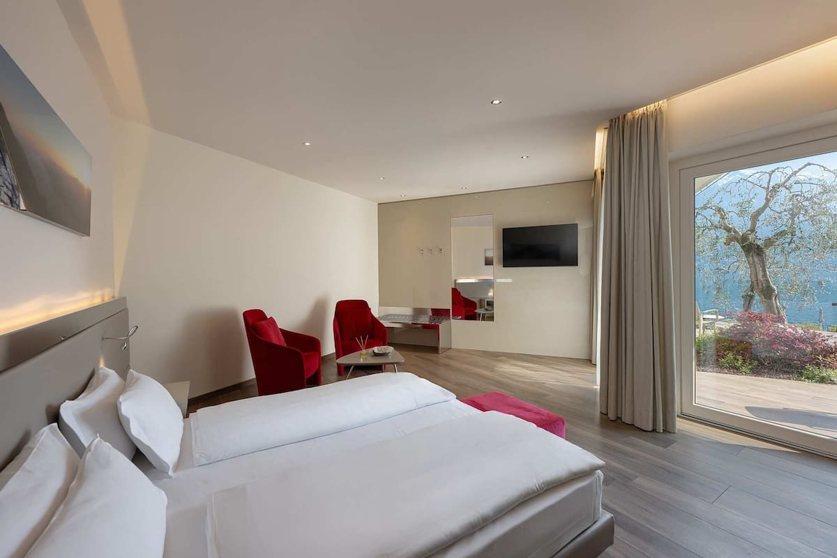 Ein modernes Hotelzimmer mit einem Bett, zwei roten Sesseln, einem kleinen Tisch, einem Fernseher und einem großen Fenster mit Blick ins Freie.