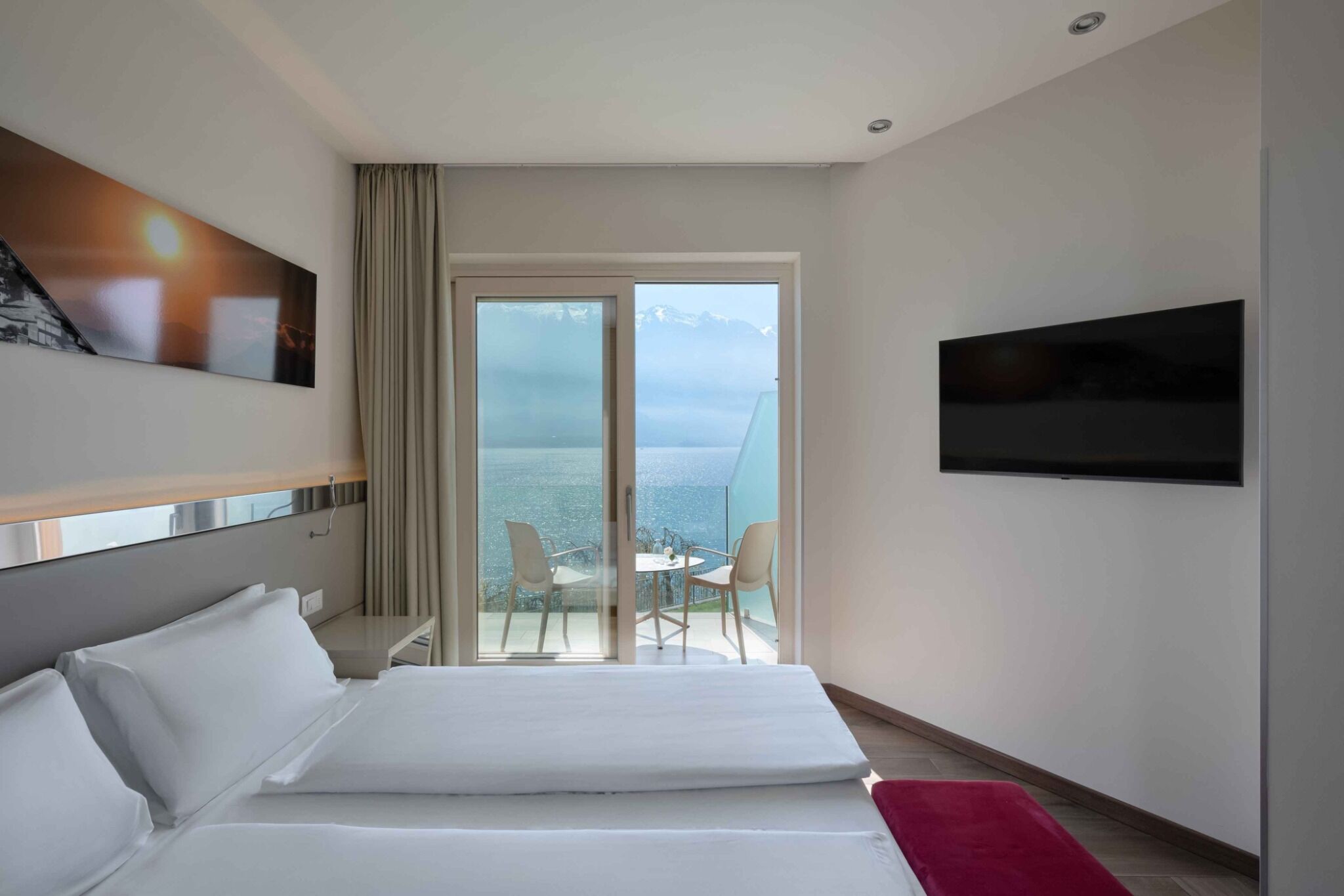 A hotel room with a bed, a TV, a balcony with a view of the sea, and a painting on the wall.