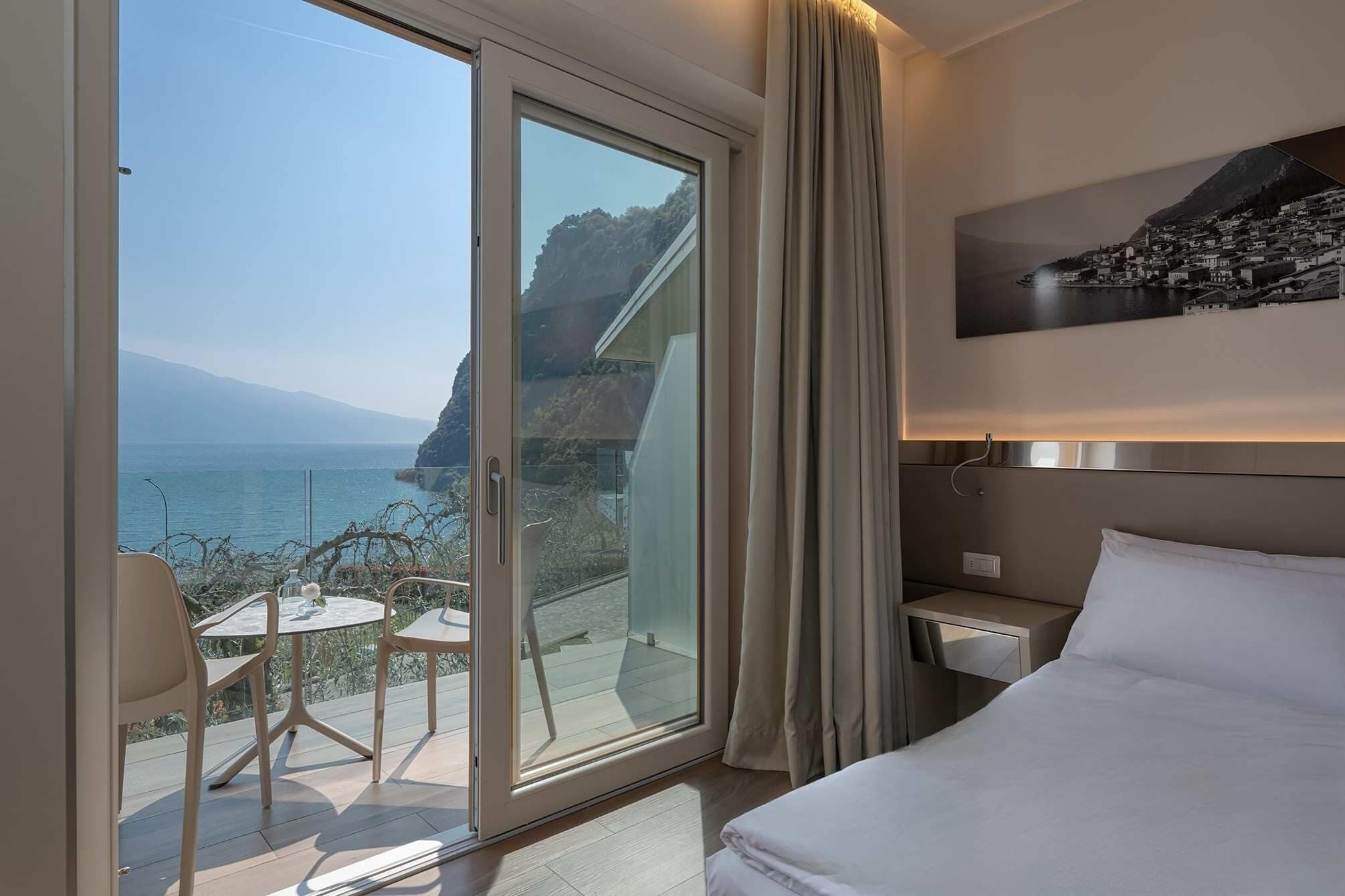 Une chambre d’hôtel avec un lit, une table de chevet, une table avec des chaises, une vue sur la mer et un tableau au mur.
