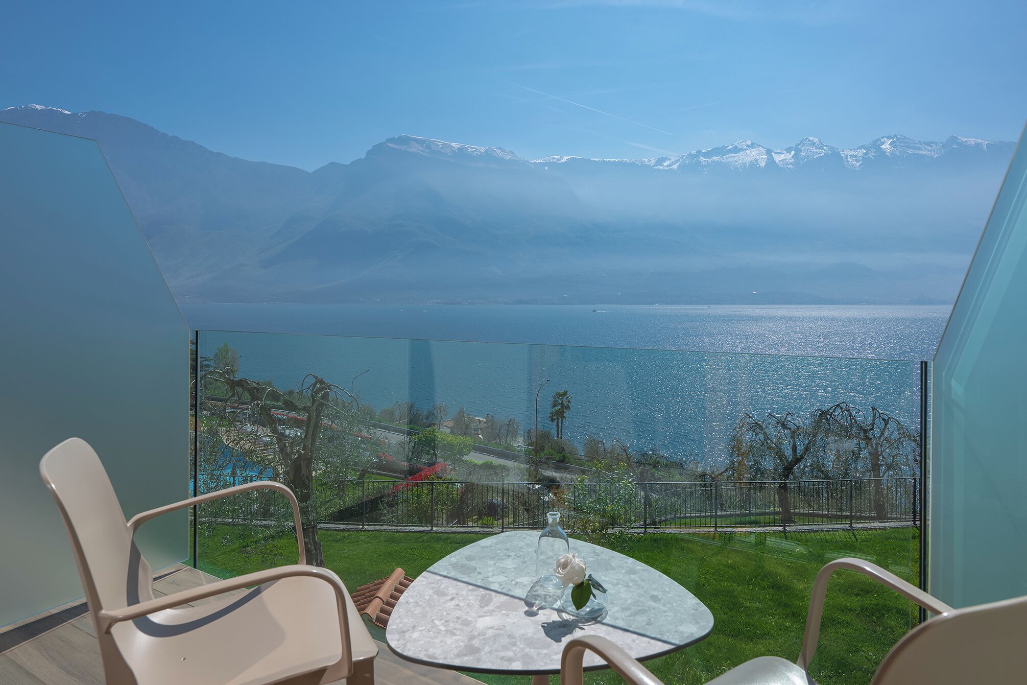 A balcony with a glass table, chairs, and a view of a mountain range and a body of water.