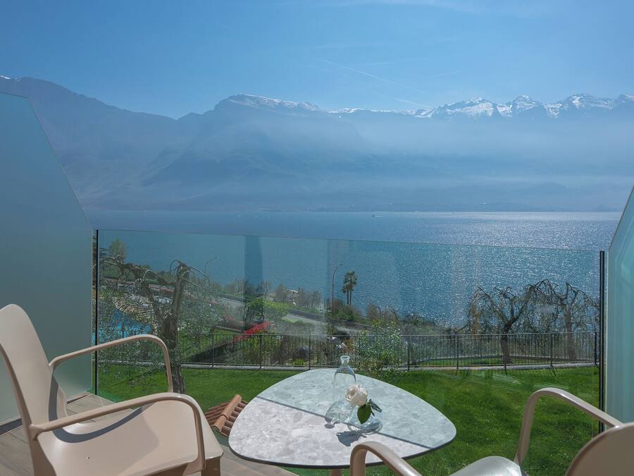 A balcony with a glass table, chairs, and a view of a mountain range and a body of water.