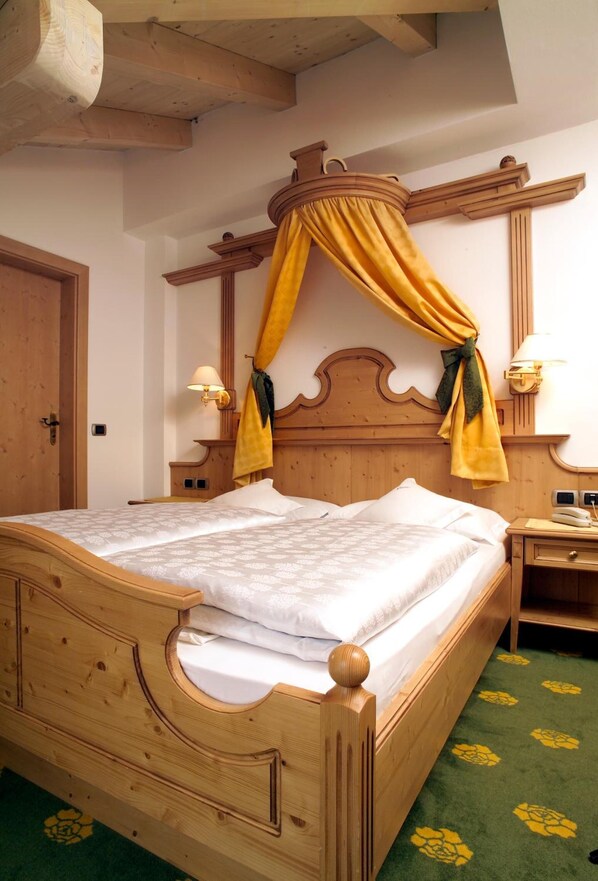A wooden four-poster bed with a canopy, a nightstand with a lamp, and a green carpet with yellow floral patterns. - Hotel Flora (Selva di Val Gardena)