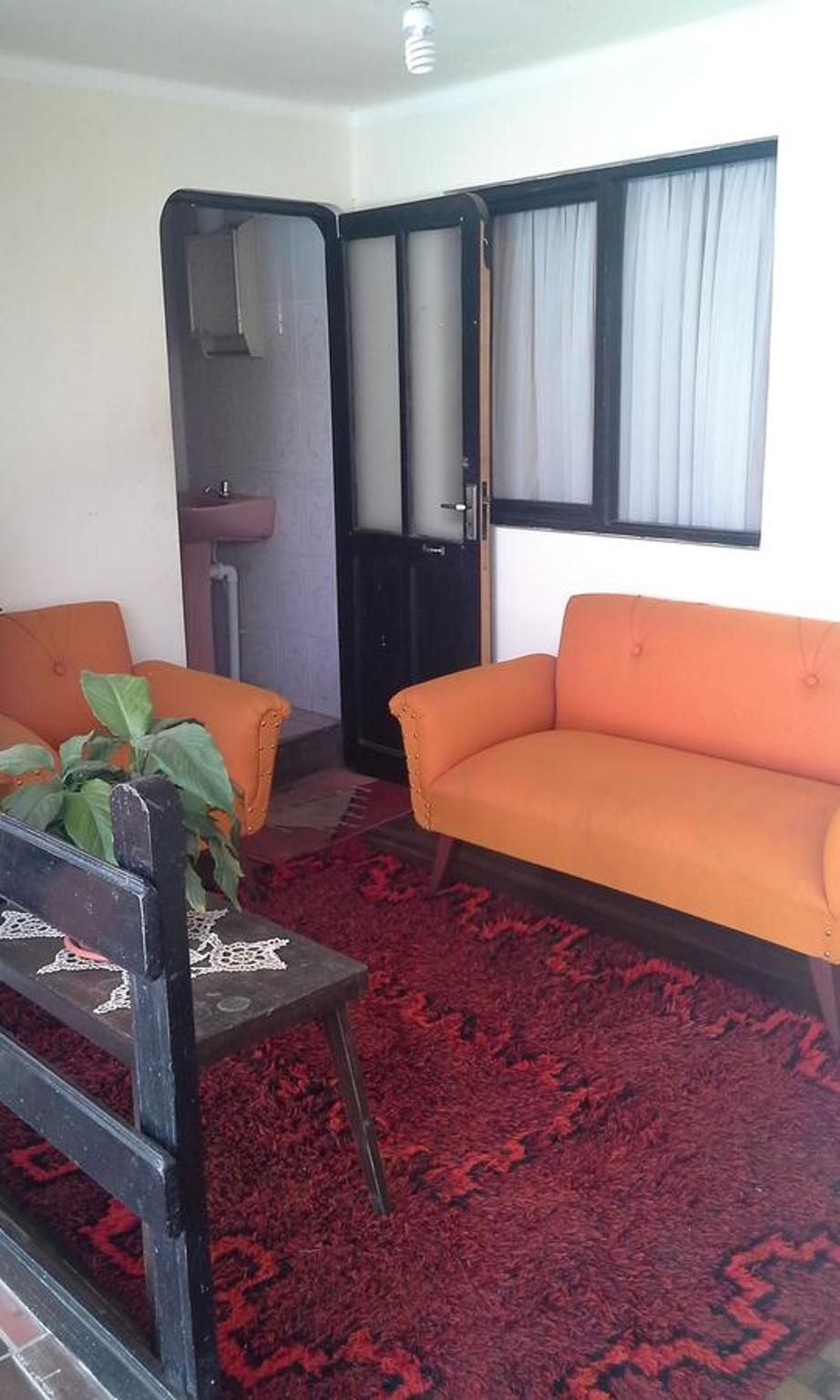 A room with a red patterned carpet, two orange sofas, a wooden chair, and a window with curtains.