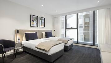 Habitación de hotel con dos camas, un sofá, una silla, una mesita y un ventanal con vistas a un edificio.