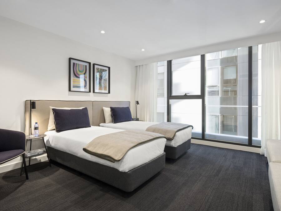 Habitación de hotel con dos camas, un sofá, una silla, una mesita y un ventanal con vistas a un edificio.