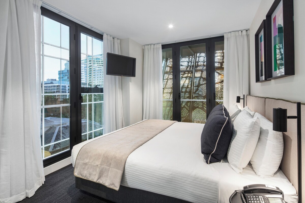 A hotel room with a large bed, a flat-screen TV, a phone, and a balcony with a view of buildings.