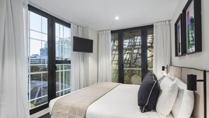 A hotel room with a large bed, a flat-screen TV, a phone, and a balcony with a view of buildings. - Quest St Kilda Road (Melbourne)