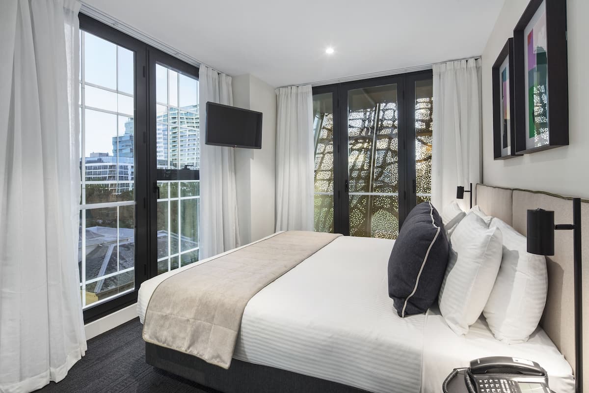 A hotel room with a large bed, a flat-screen TV, a phone, and a balcony with a view of buildings.