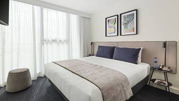 A modern hotel room with a large bed, a bedside table with a phone and water bottle, a small round ottoman, and a TV mounted on the wall.