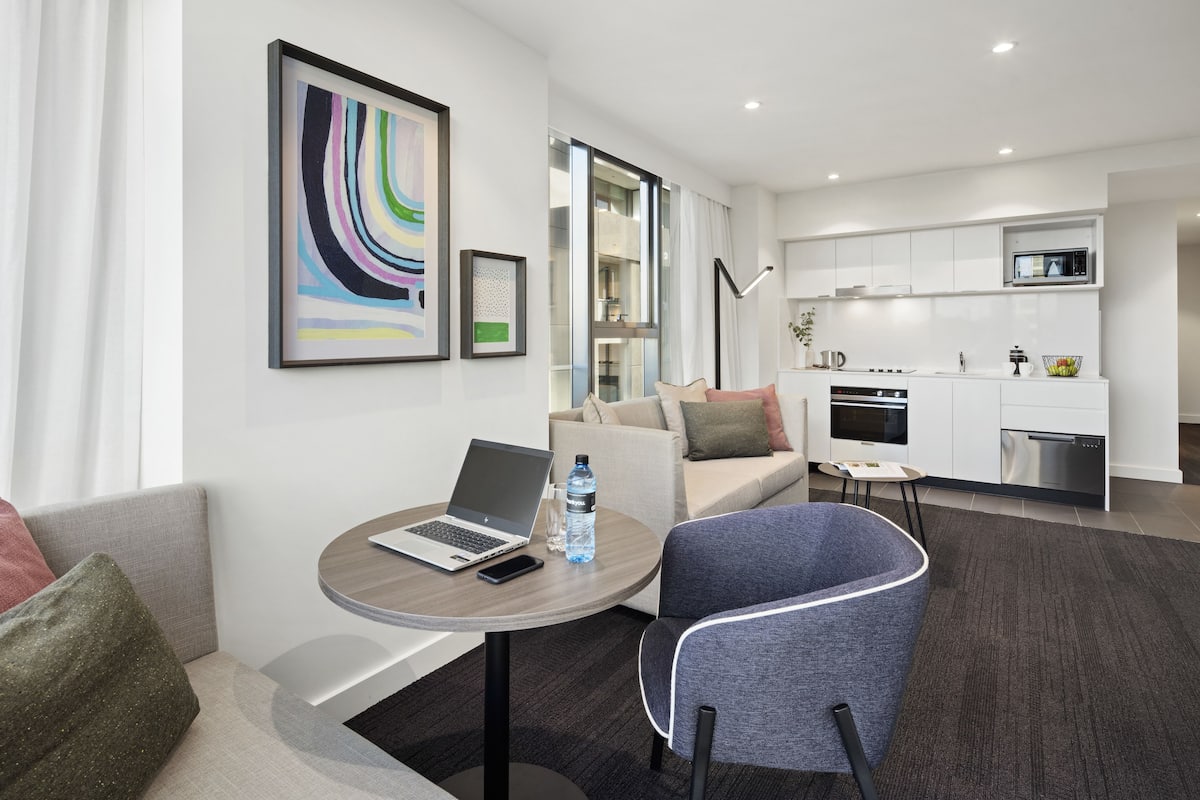 A modern hotel room with a sofa, a round table with a laptop, a chair, a kitchen area with a microwave, oven, and dishwasher.