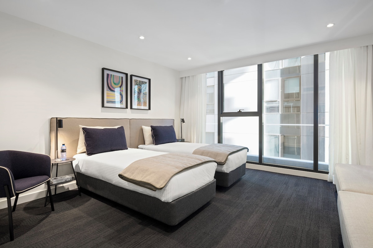A hotel room with two beds, a sofa, a chair, a small table, and a large window with a view of a building.