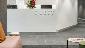 A modern reception area with a white counter, a flower arrangement, and a seating area with a couch and a small table. - Quest St Kilda Road (Melbourne)