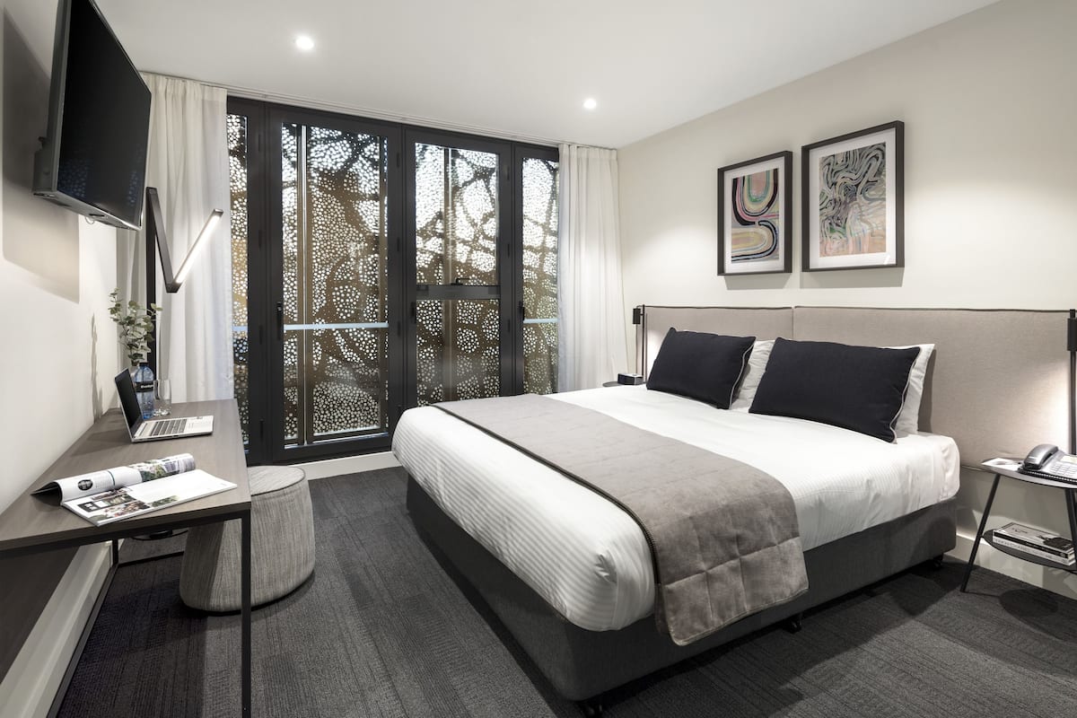 A modern hotel room with a large bed, a desk with a laptop, a flat-screen TV, and artwork on the walls.