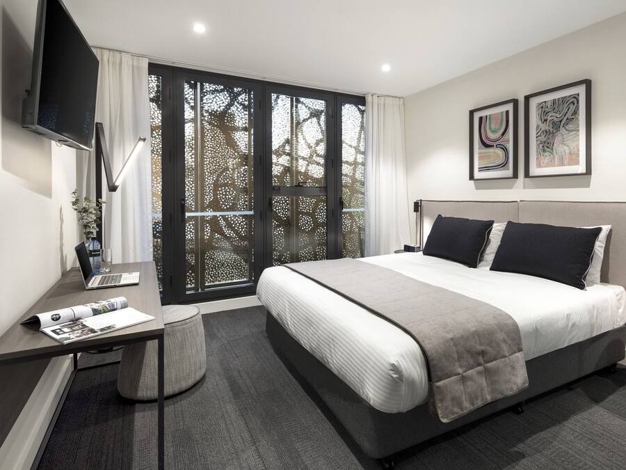 Una habitación de hotel moderna con una cama grande, un escritorio con una computadora portátil, un televisor de pantalla plana y obras de arte en las paredes.