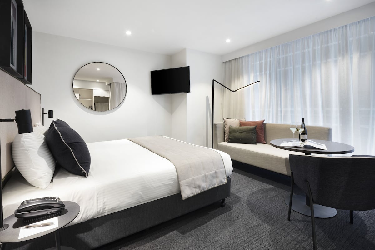 A modern hotel room with a bed, a sofa, a dining table, a TV, and a large window with curtains.