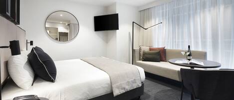 Ein modernes Hotelzimmer mit Bett, Sofa, Esstisch, Fernseher und einem großen Fenster mit Vorhängen.