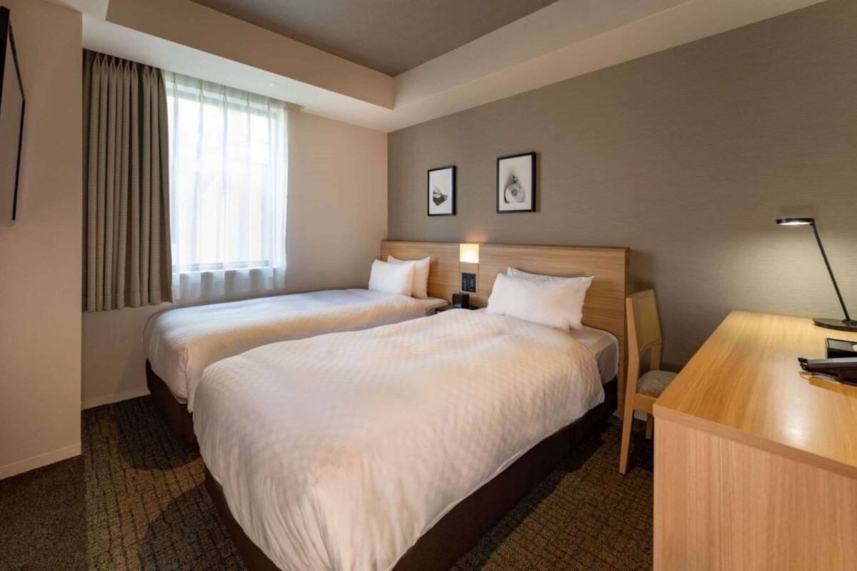 Habitación de hotel con dos camas, un escritorio, una lámpara y una ventana con cortinas.