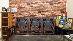 Una habitación con pared de ladrillo, un mueble de madera con cajones, un espejo decorativo con centro verde y una pequeña mesa con varios objetos.