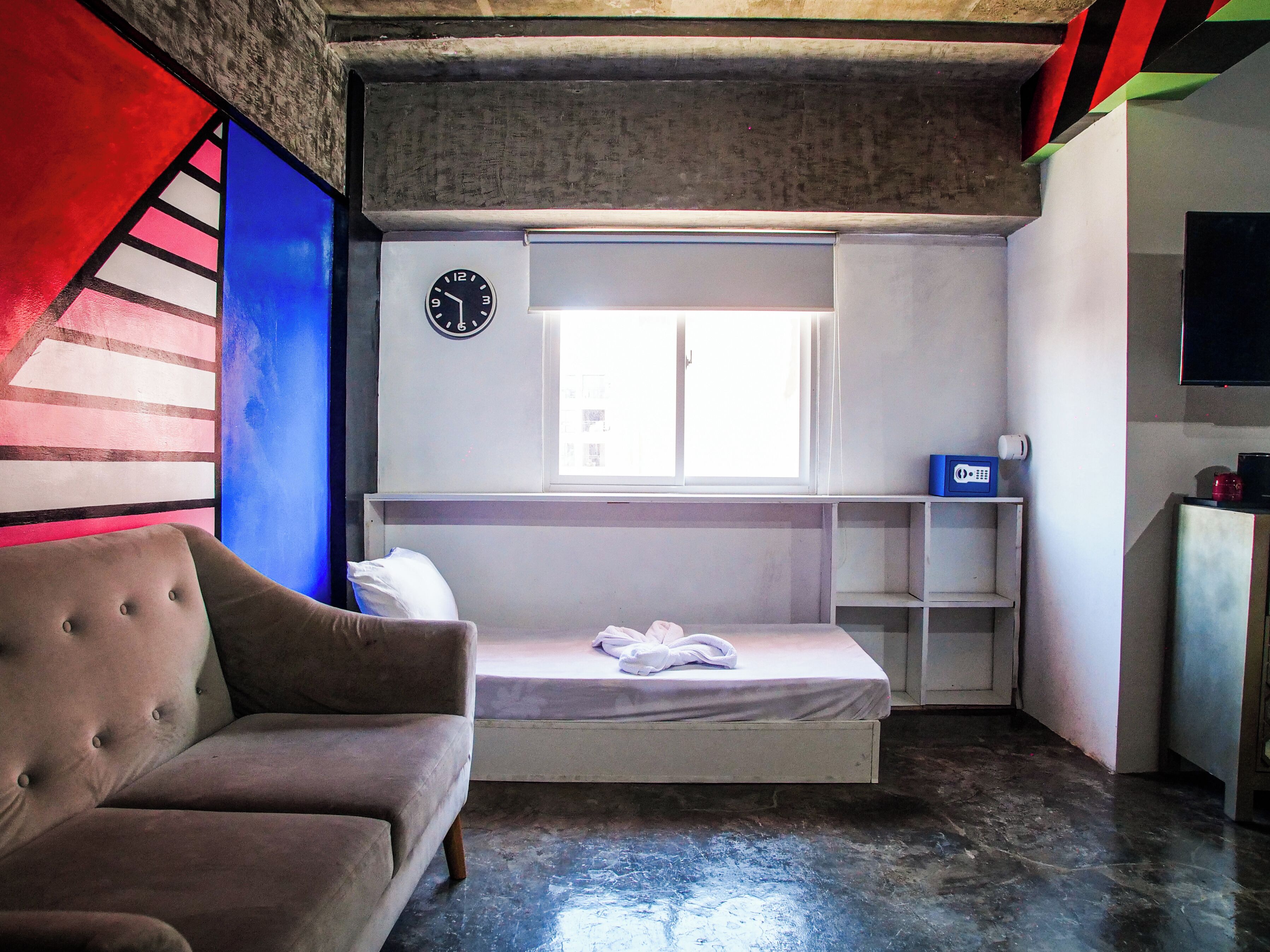 Una habitación pequeña y funcional con una cama individual, un sofá, un reloj en la pared y una ventana que proporciona luz natural.