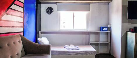 Una habitación pequeña y funcional con una cama individual, un sofá, un reloj en la pared y una ventana que proporciona luz natural.