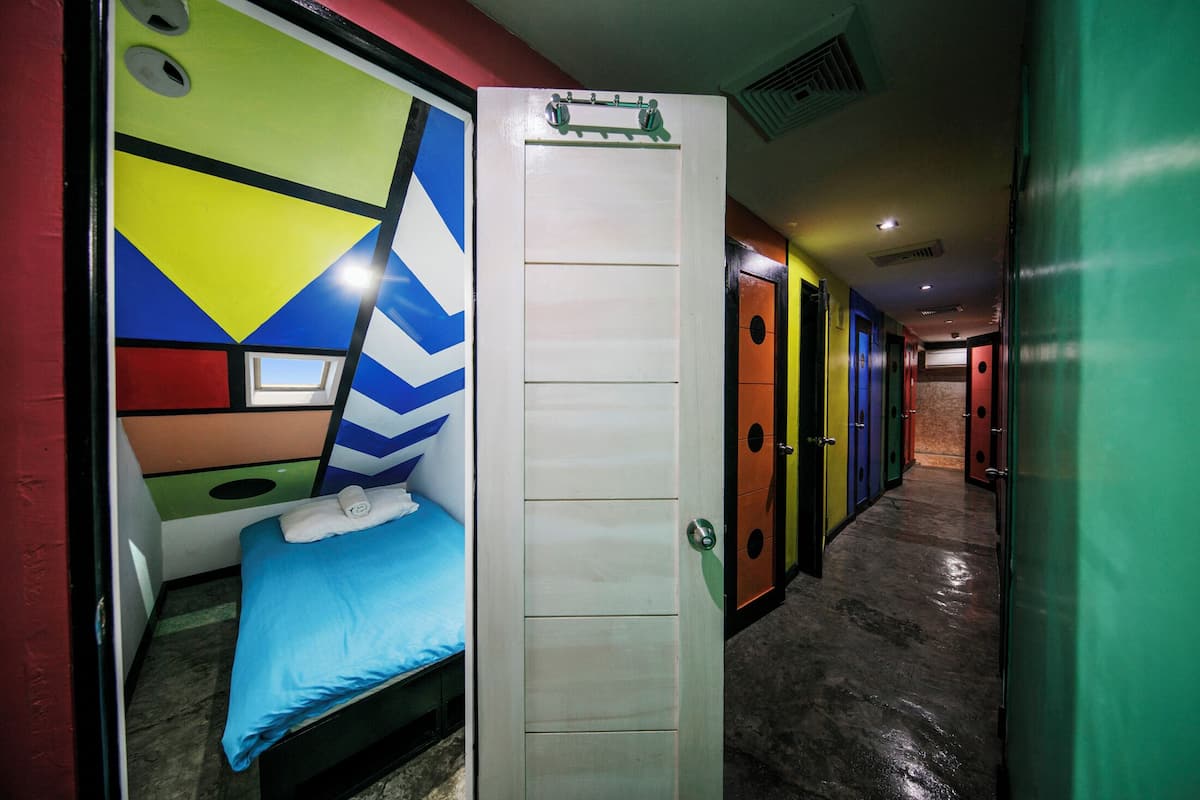 A hotel room with a bed, a window, and a door leading to a hallway with colorful doors.