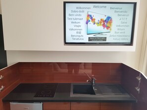 A modern kitchen with a built-in stove, a sink, and a large flat-screen TV displaying a welcome message in multiple languages. - Best Western Hotel Journel Paris Sud (Ris Orangis)