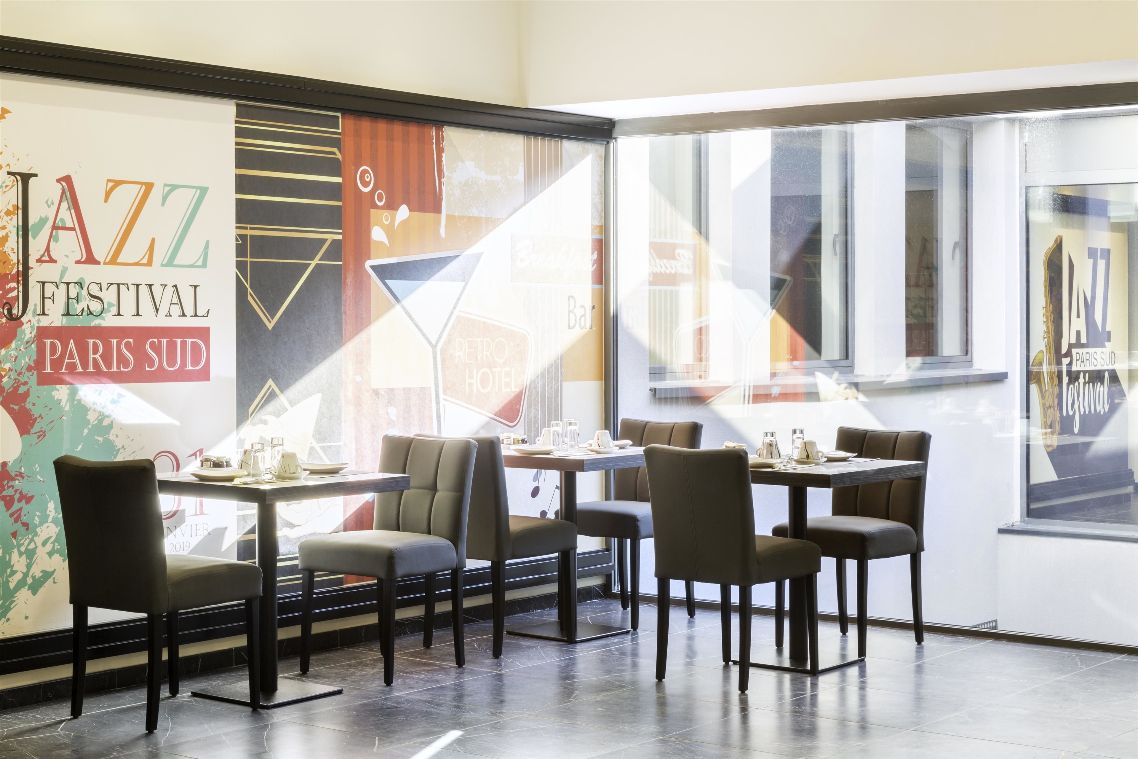 A restaurant with a retro theme, featuring a large poster of the Jazz Festival Paris Sud, multiple tables and chairs, and a view of the street outside.