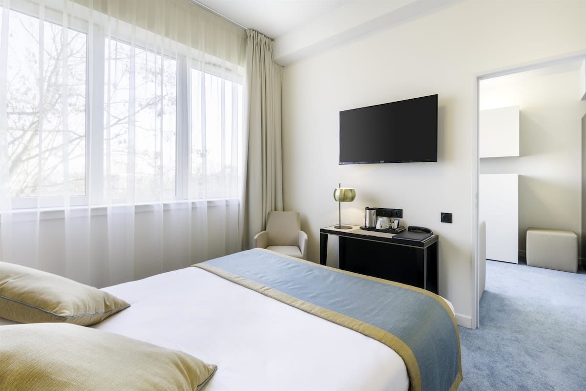 A hotel room with a large bed, a TV mounted on the wall, a desk with a lamp, and a view of trees through the window.