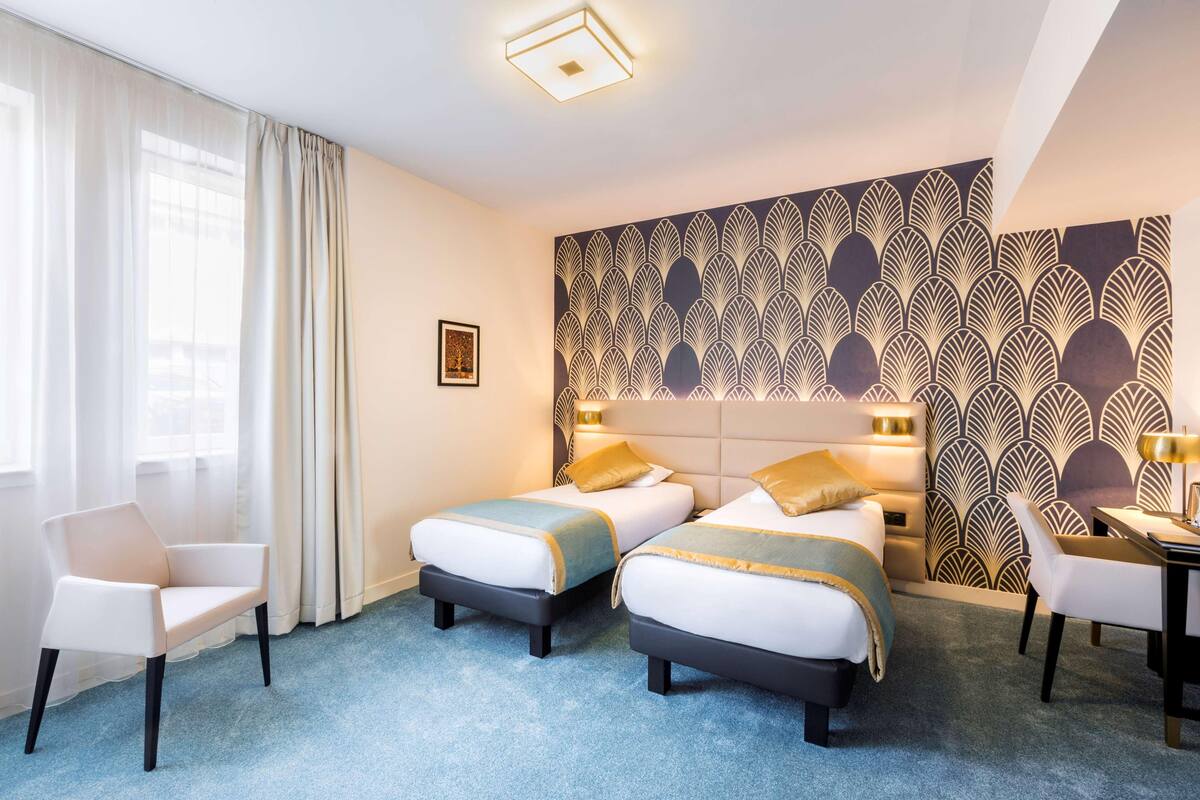 A hotel room with two beds, a chair, a desk, and a patterned wall.