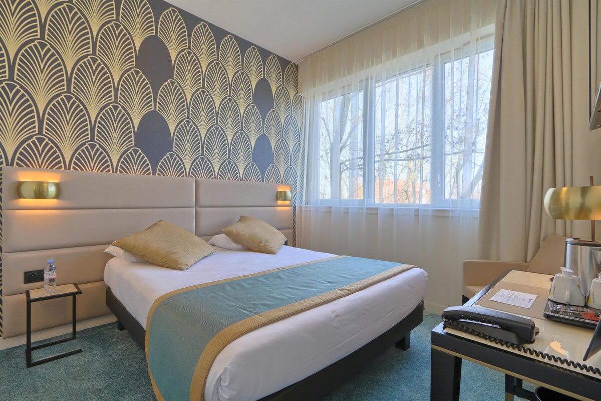A hotel room with a large bed, a bedside table with a lamp, a desk with a telephone, and a window with curtains.