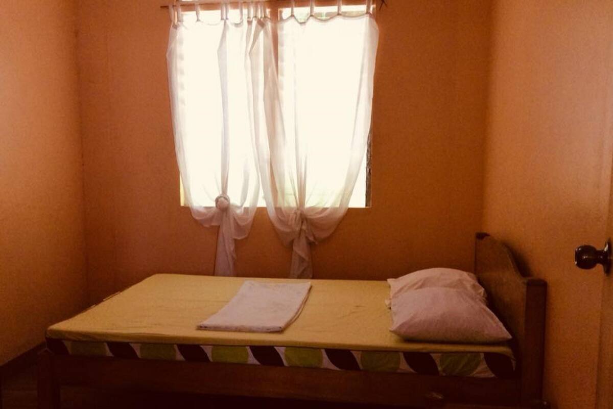 A single bed with a wooden headboard, a bedside table, and a window with sheer curtains tied back.