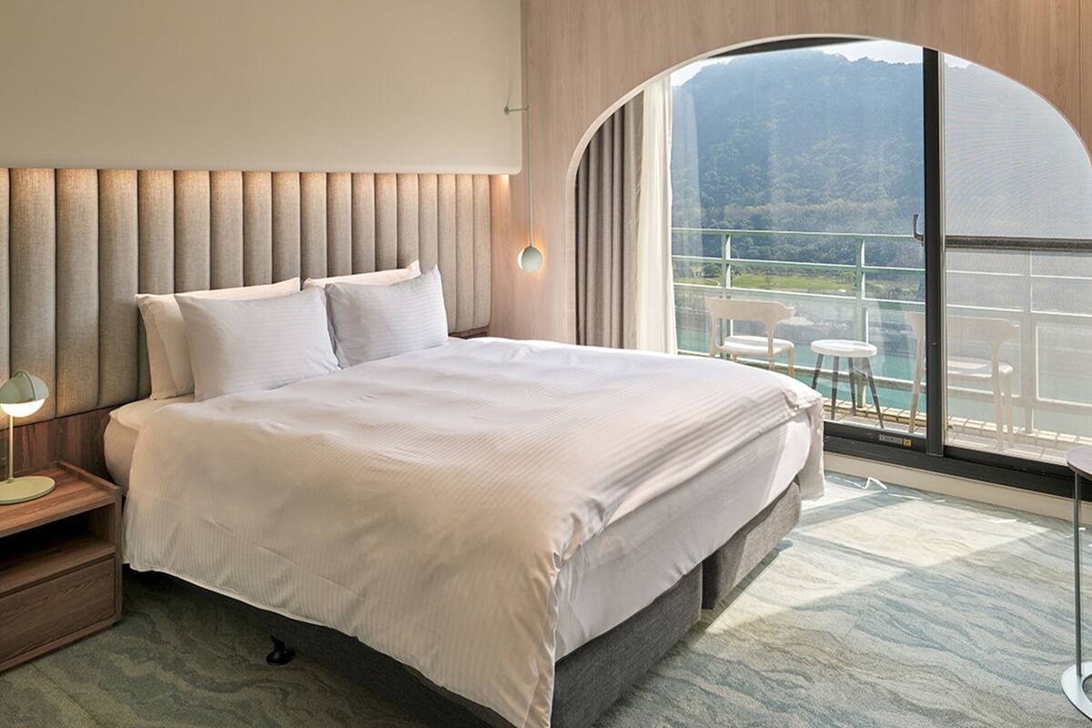 A modern hotel room with a large bed, a nightstand with a lamp, a small table, and a view of a balcony with outdoor seating.