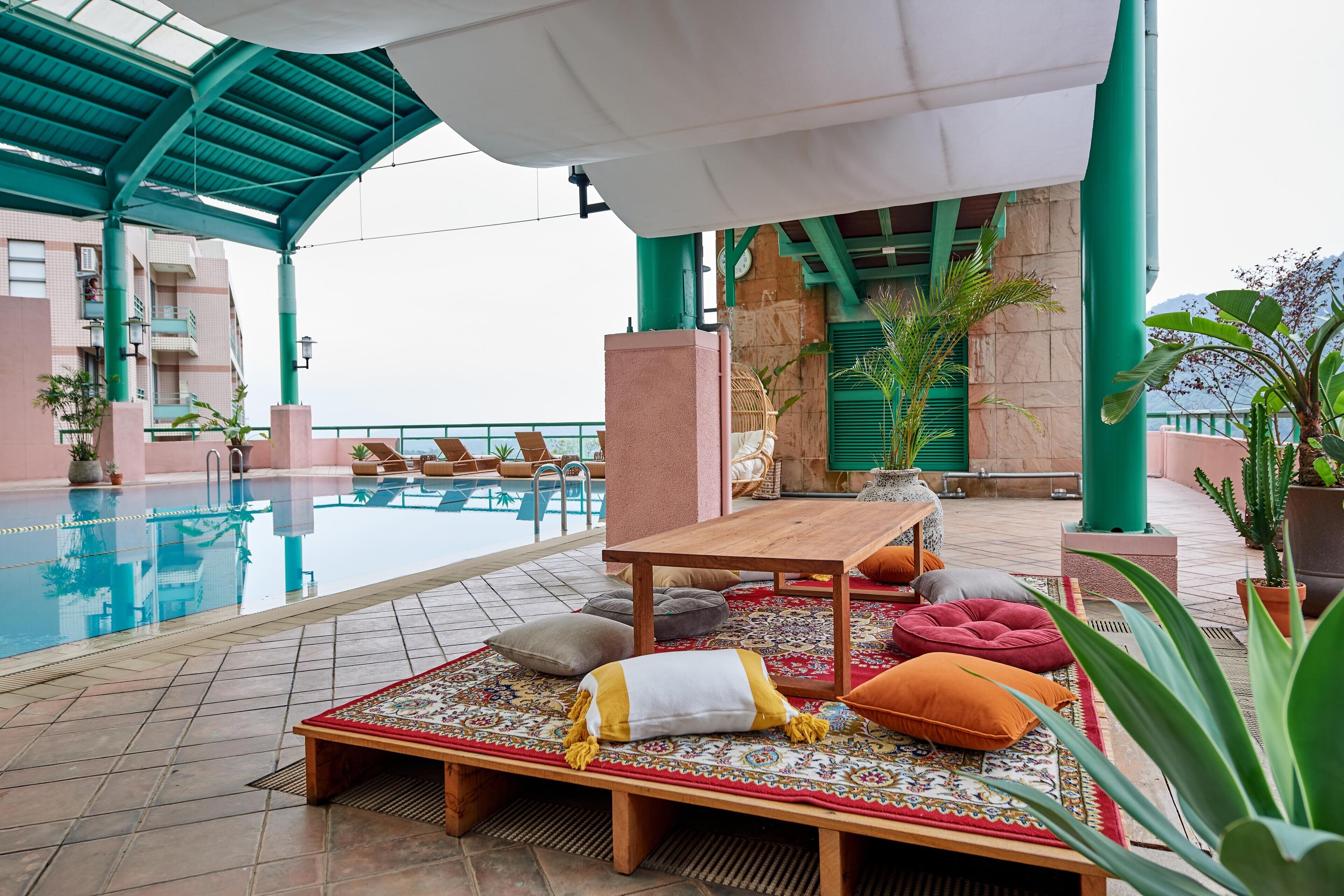 A poolside area with a wooden bench, cushions, and a patterned rug.