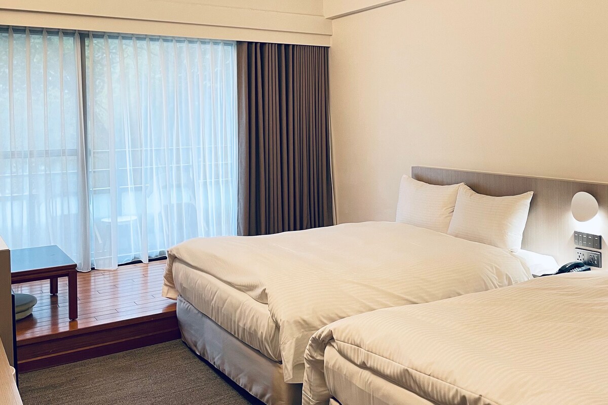 A hotel room with two beds, a desk, and a view of the outdoors.