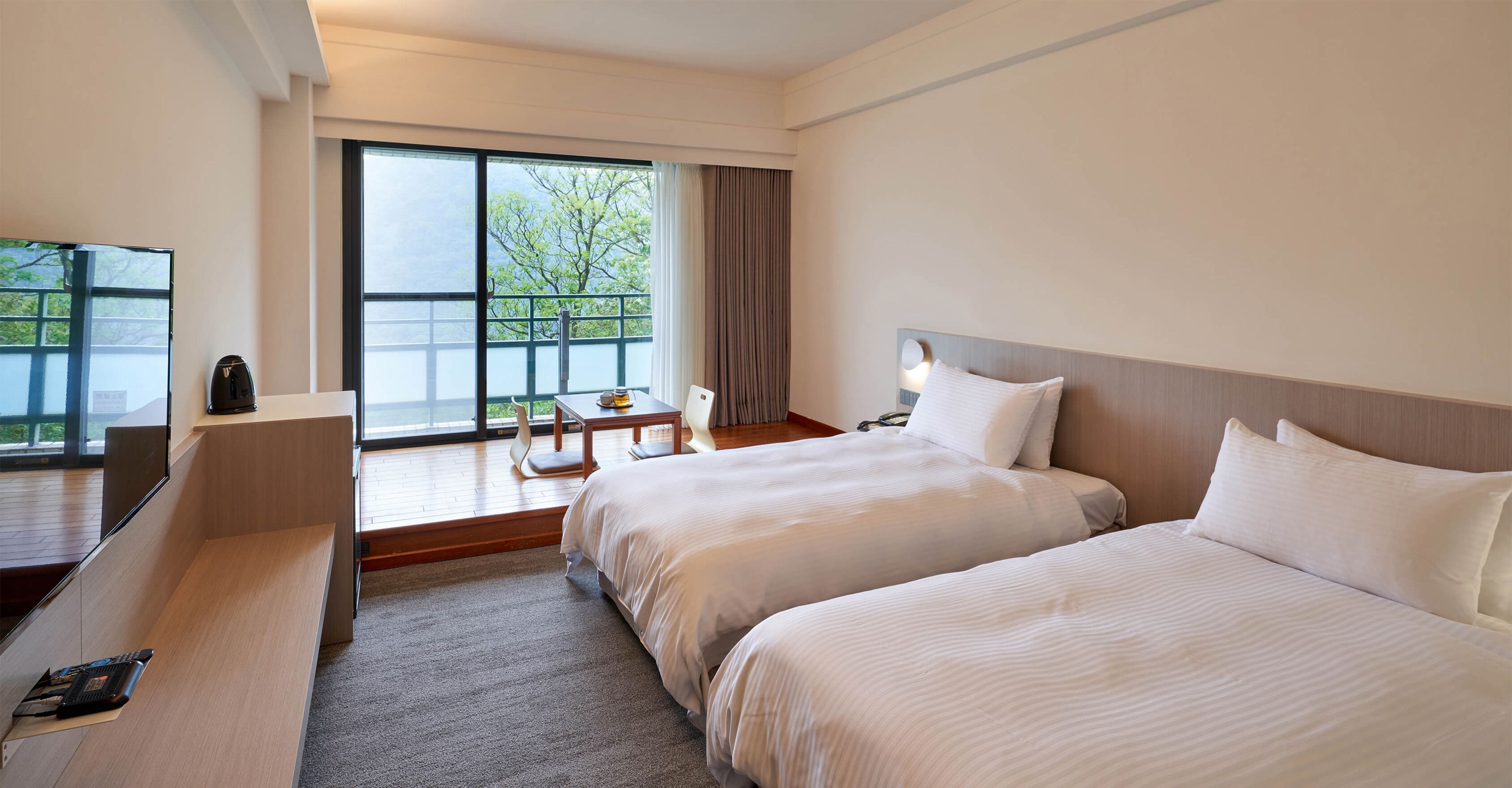 A hotel room with two beds, a desk, a chair, a TV, and a window with a view of trees.