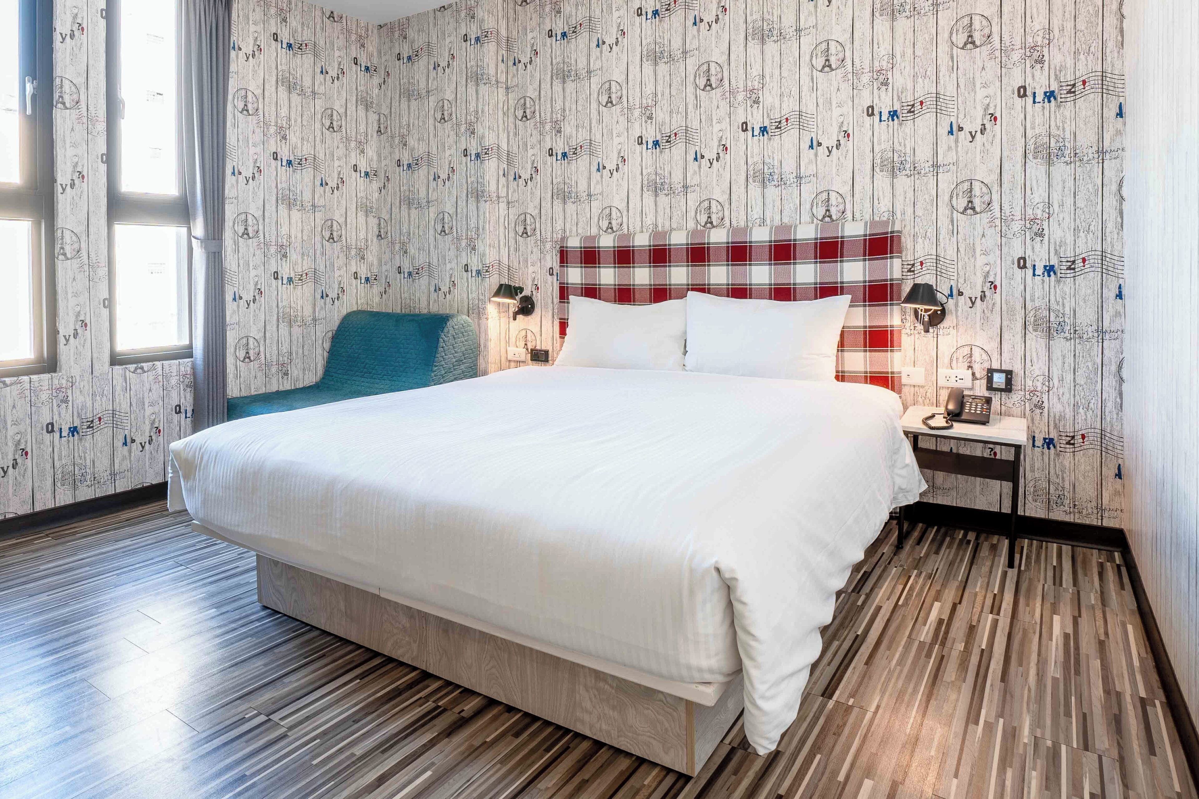 A neatly made bed with a plaid headboard, a bedside lamp, and a small table with a phone. The room has a wooden floor and a patterned wallpaper.