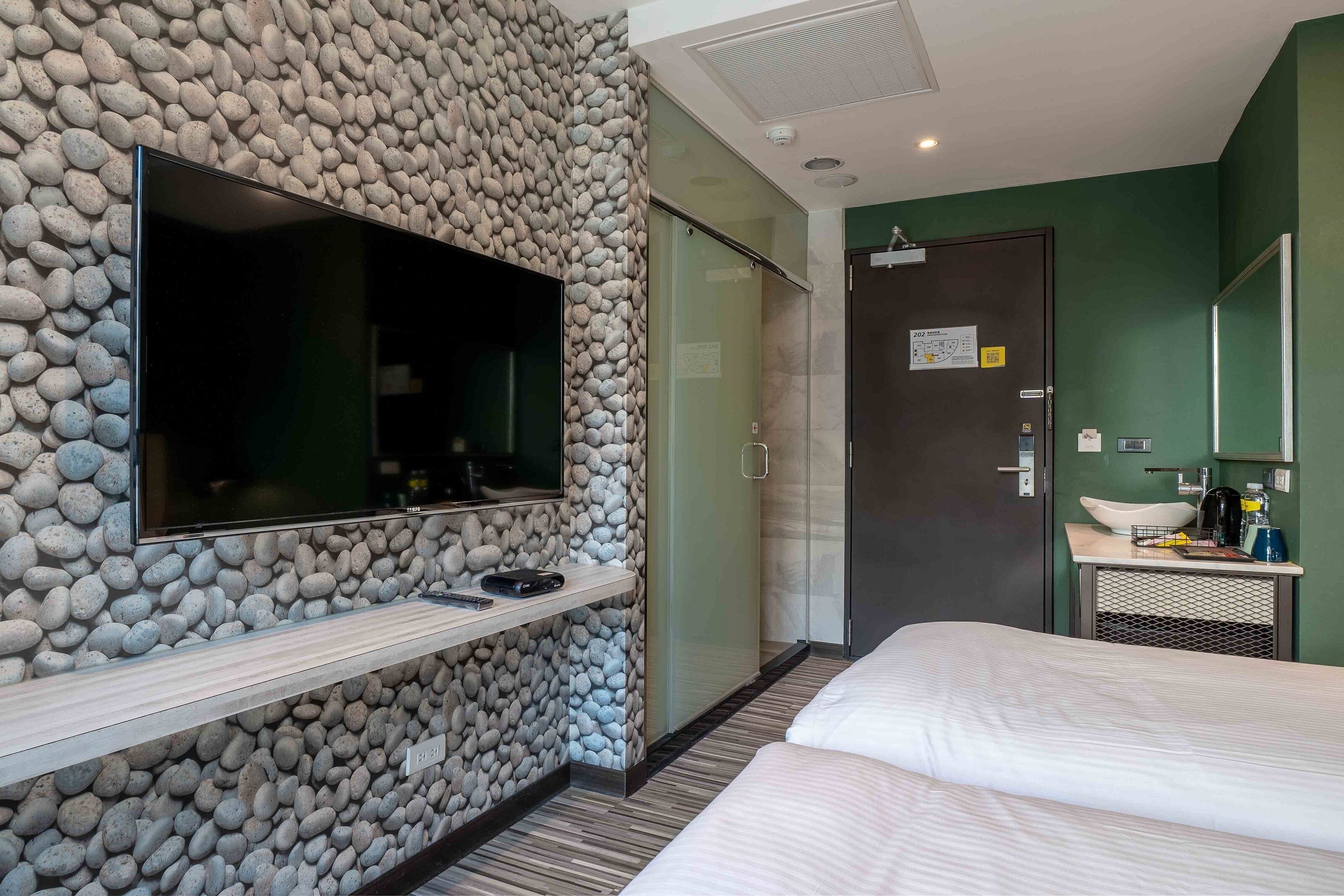 A hotel room with a stone wall, a flat-screen TV, a bed, a bathroom with a sink and mirror, and a door with a sign.