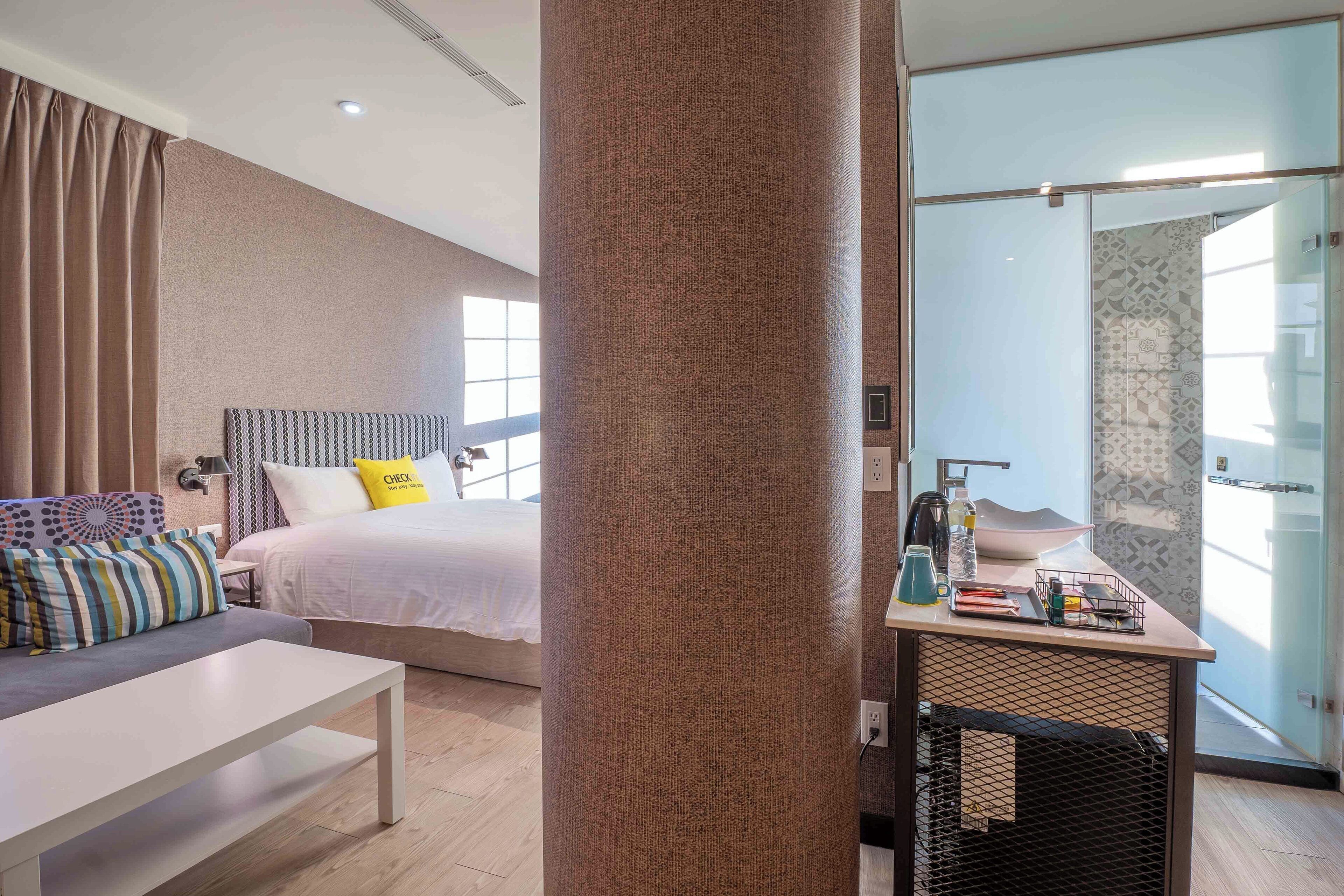 Ein modernes Hotelzimmer mit einem Bett, einer Couch, einem Schreibtisch mit Wasserkocher und einer Glaskonstruktion.
