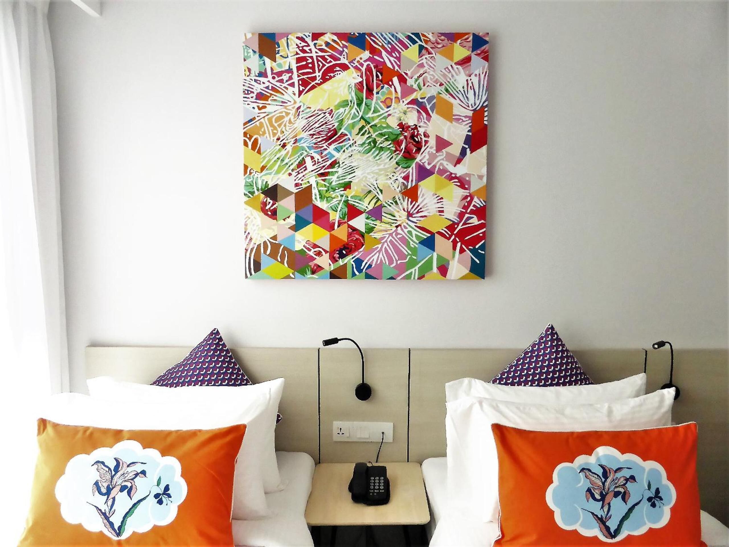 Une chambre d’hôtel avec un lit, deux oreillers ornés de motifs floraux, une table de chevet avec un téléphone et une peinture abstraite aux couleurs vives accrochée au mur.